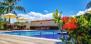 Una piscina amb gandules i ombrel·les, envoltada de palmeres i un edifici amb teulada de rajola.