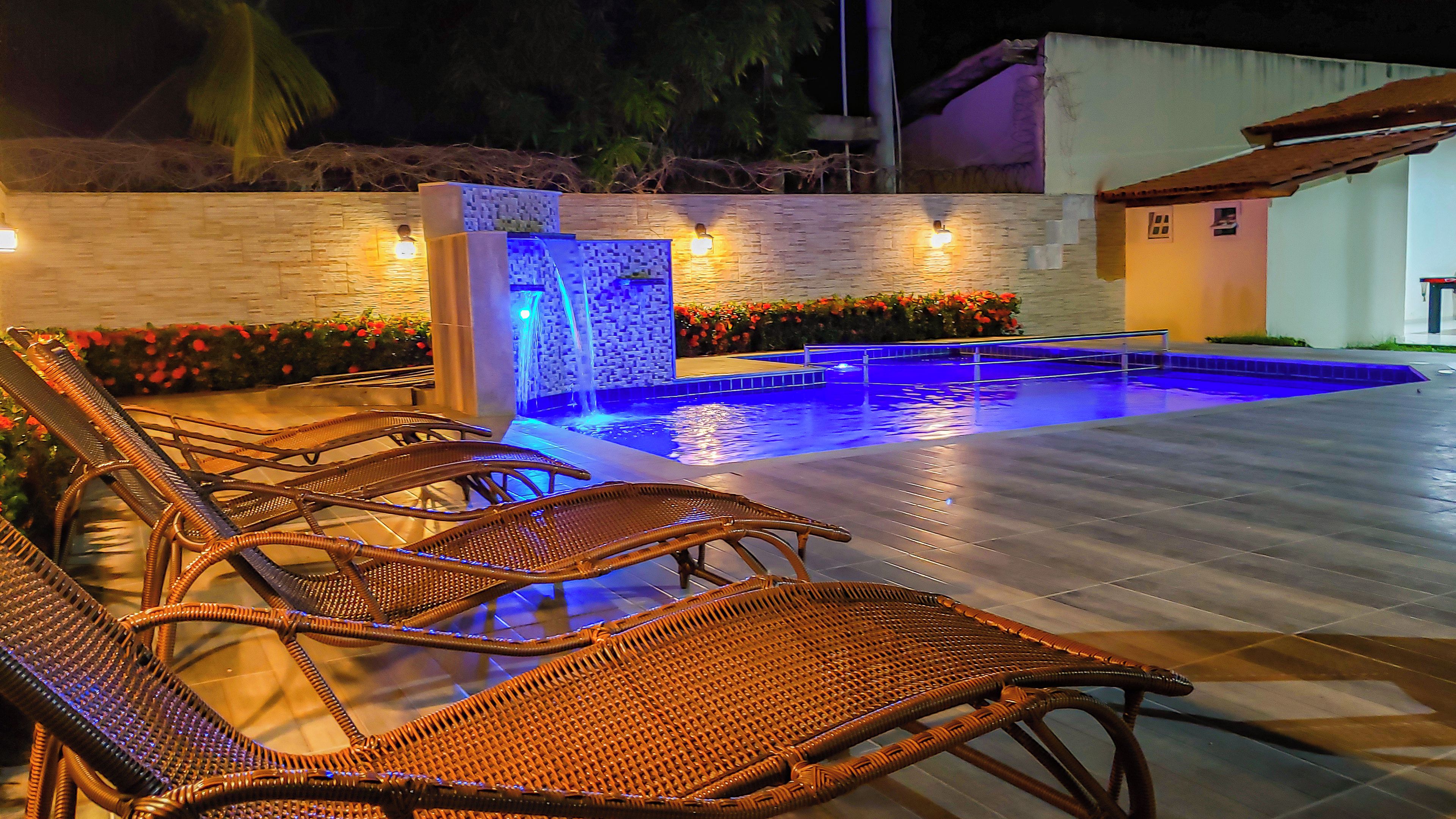 Piscina exterior com efeito cascata, rodeada por cadeiras de vime e um muro de pedra.
