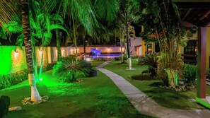 A well-lit outdoor area with a pathway leading to a pool area, surrounded by various tropical plants and palm trees. - Bem Bahia Hotel (Santa Cruz Cabralia)
