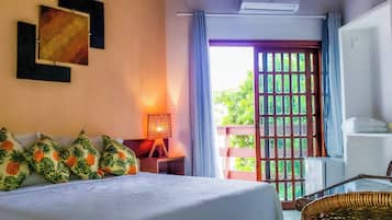 A hotel room with a bed, a nightstand with a lamp, a wicker chair, and a view of greenery through a window.