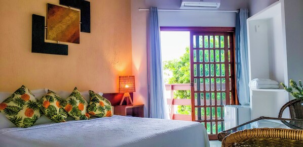 Habitación de hotel con una cama, una mesita de noche con lámpara, un sillón de mimbre y vistas a la vegetación por la ventana.