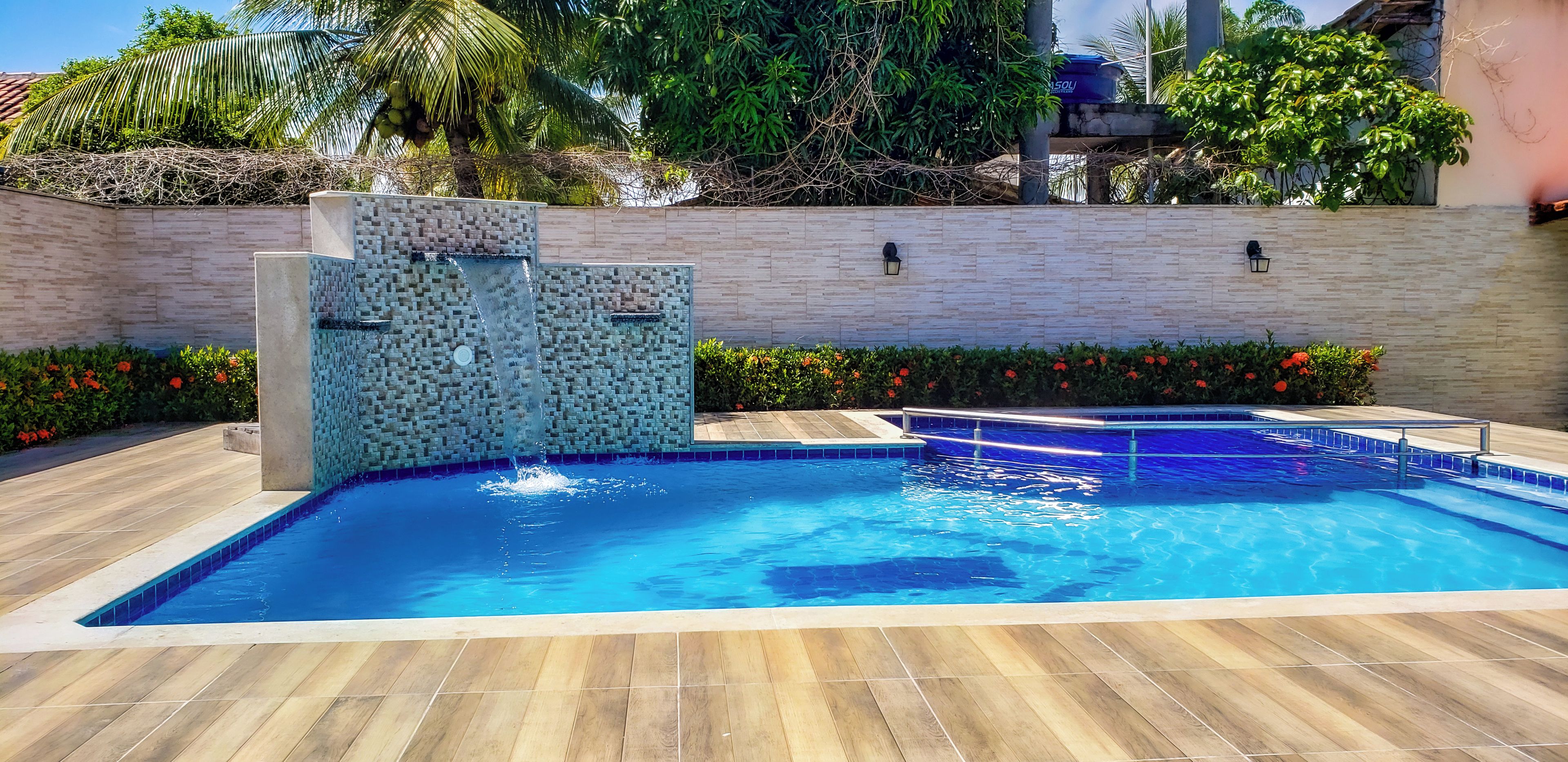 Una piscina con una cascada de azulejos, rodeada por una terraza de madera y un bajo muro de ladrillo con plantas.
