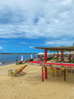 Uma praia com um toldo identificado como 'JESUS CRISTO MESTRE' e cadeiras de praia.
