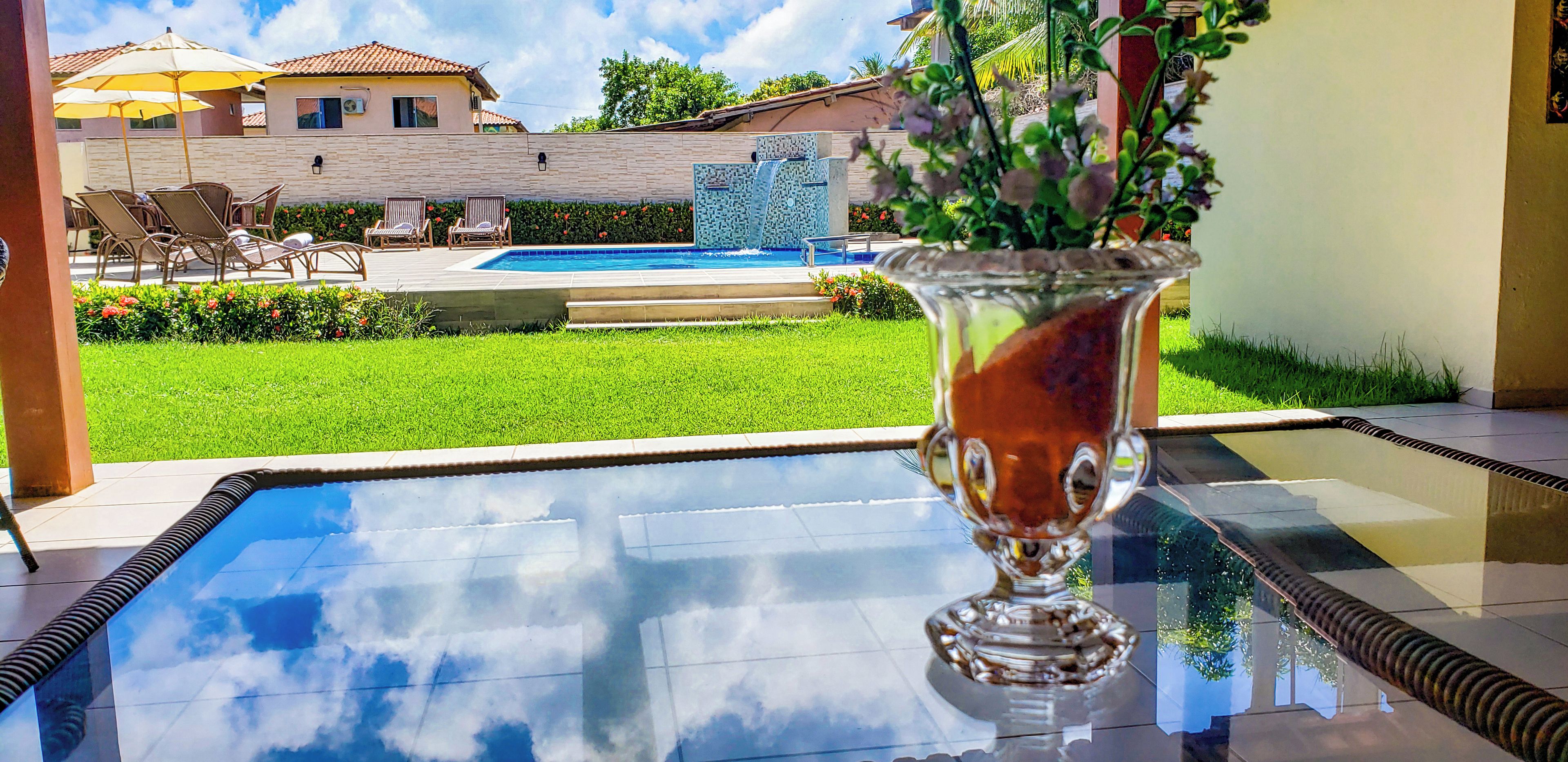 A glass-top pool table with a vase of flowers, outdoor seating, and a view of a swimming pool and buildings in the background.