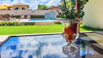 A glass-top pool table with a vase of flowers, outdoor seating, and a view of a swimming pool and buildings in the background.