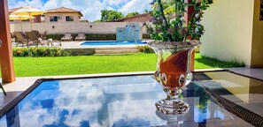 Una mesa de pool con superficie de vidrio, un jarrón de flores, mobiliario exterior y vistas a una piscina y edificios al fondo.