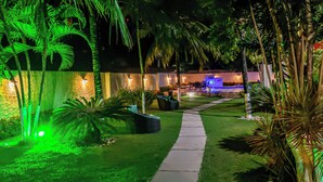 Un jardín nocturno con un sendero, palmeras y un elemento acuático.