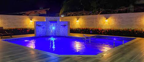 A swimming pool with a blue-lit water feature, surrounded by a stone wall and red flowers.