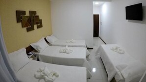 A hotel room with four beds, each with a white bedspread and a towel on the bed. There is a flat-screen TV mounted on the wall and a door leading to another room.