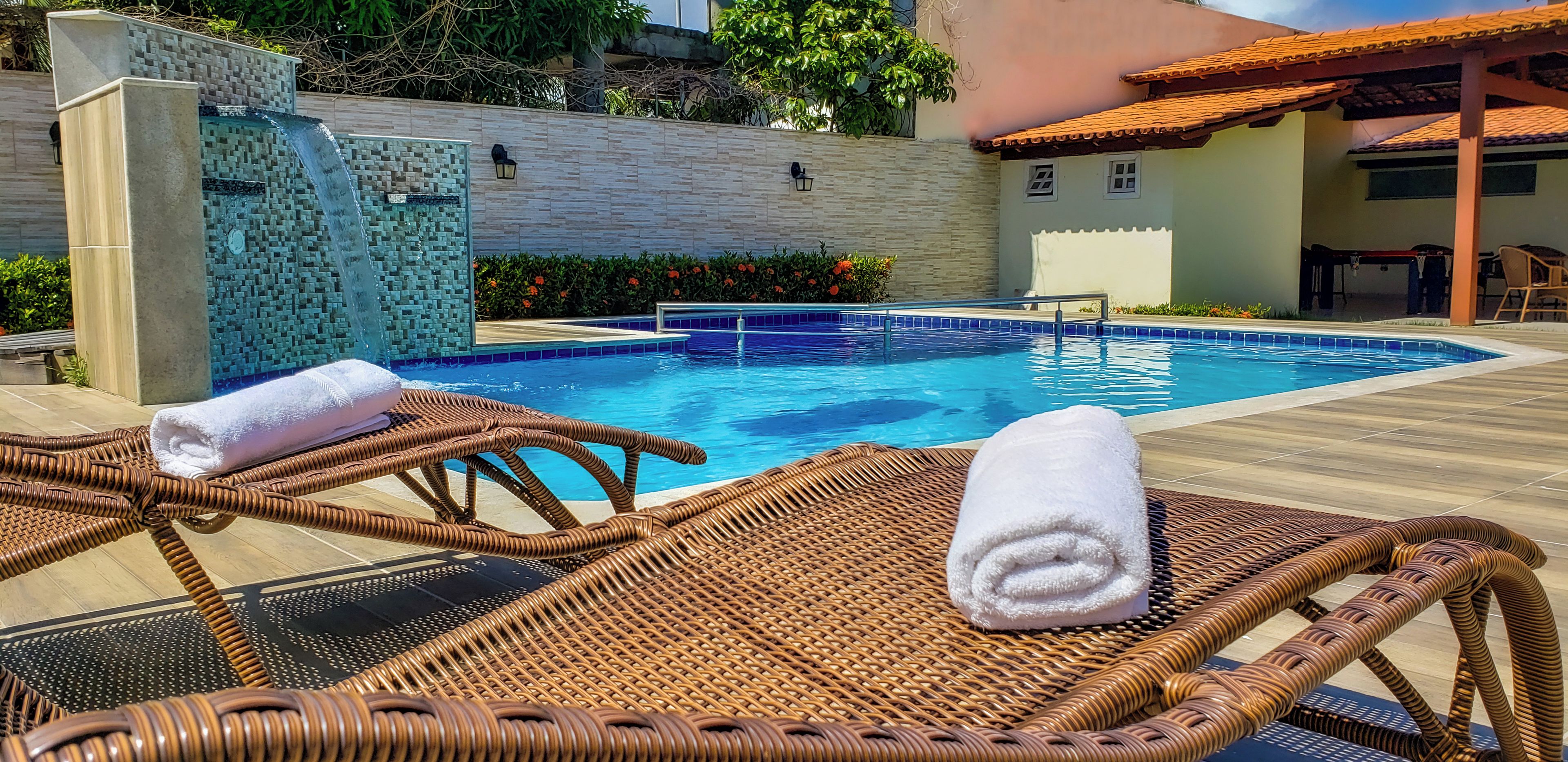 Una piscina con un muro de mosaico, sillones de mimbre con toallas y una zona con techo de baldosas.