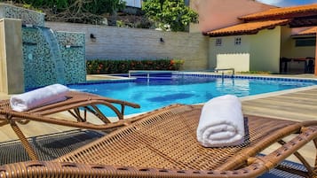 Una piscina con un muro de mosaico, sillones de mimbre con toallas y una zona con techo de baldosas.