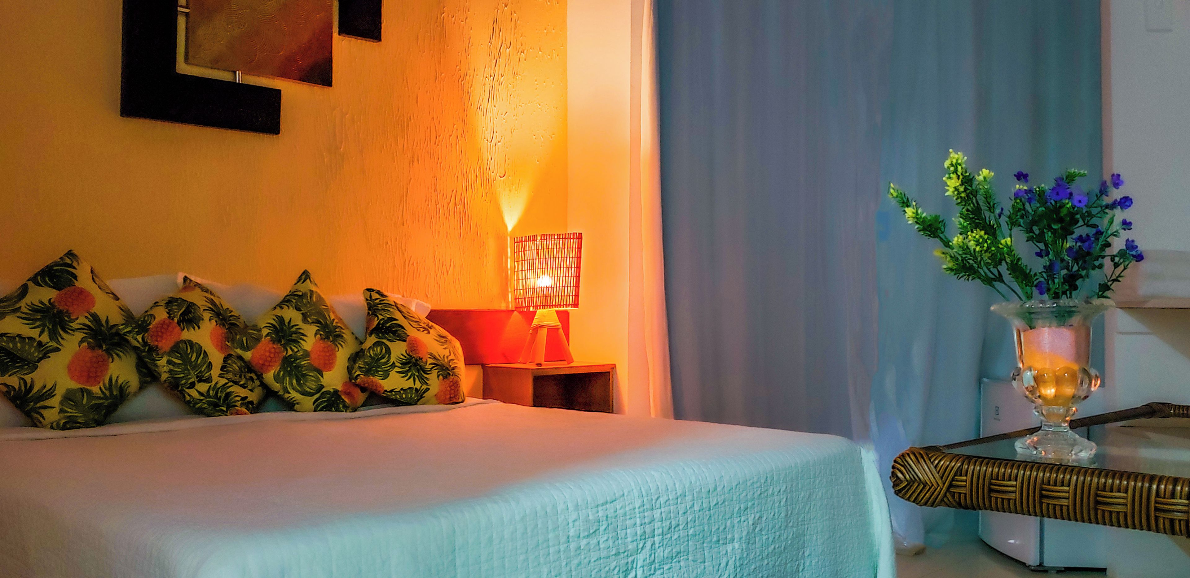 A hotel room with a bed, tropical-themed pillows, a nightstand with a lamp, and a vase with flowers.