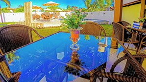 A glass-top table with wicker chairs, a potted plant, and a view of a pool and outdoor seating area.