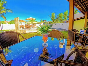 A glass-top table with wicker chairs, a potted plant, and a view of a pool and outdoor seating area. - Bem Bahia Hotel (Santa Cruz Cabralia)