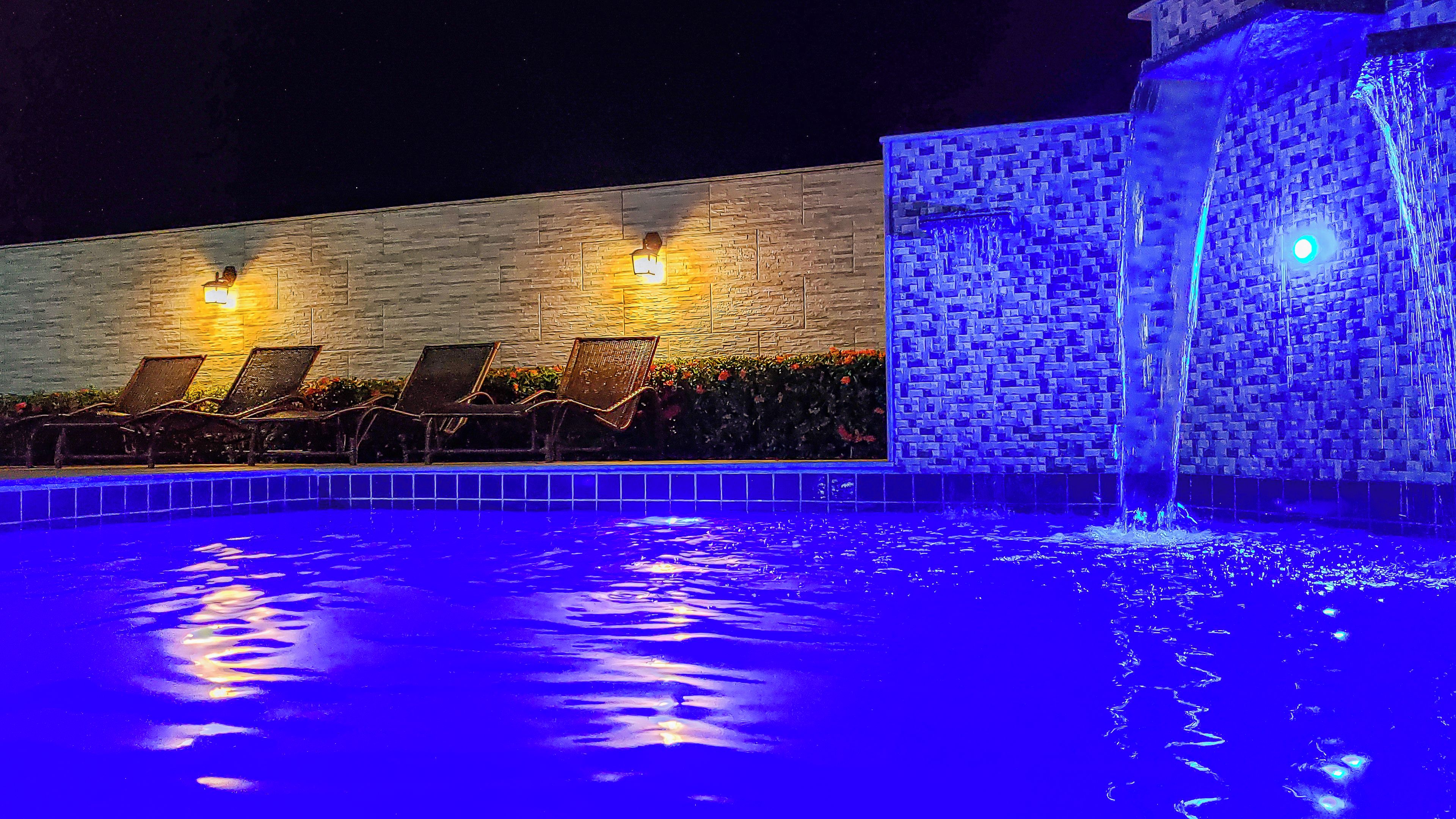 Ein Poolbereich bei Nacht mit Liegestühlen und einem Wasserspiel.