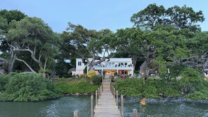 A wooden pier leading to a white building with a balcony, surrounded by dense greenery and a calm body of water.