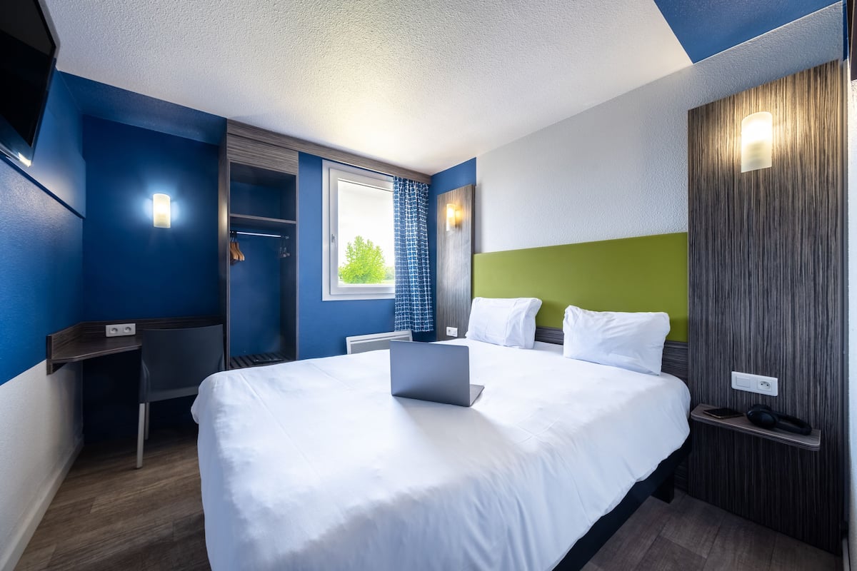 A hotel room with a bed, a desk, a chair, a window with curtains, and a wall-mounted light.