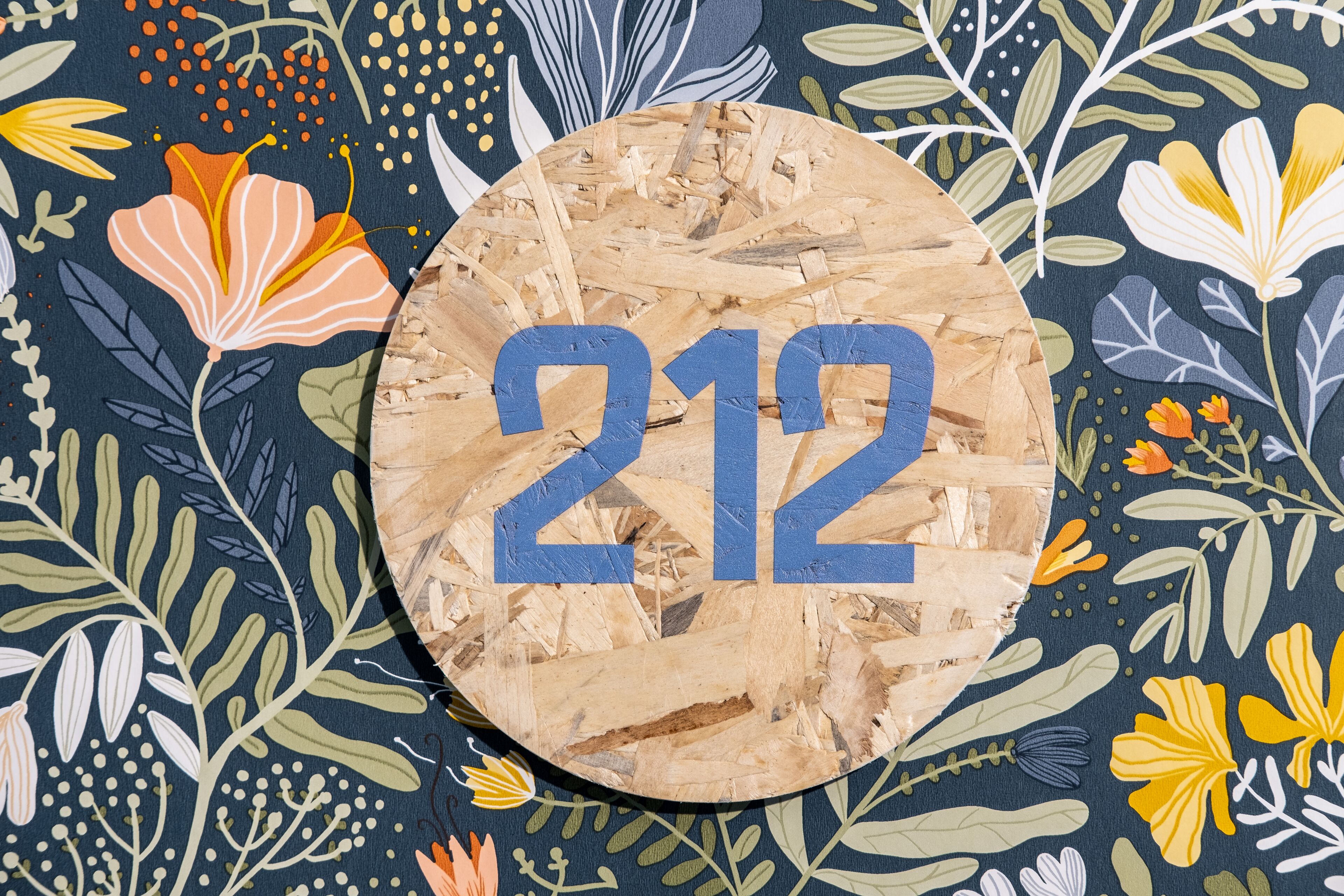 Een rond houten bordje met het nummer 212 op een achtergrond met bloemenmotief.