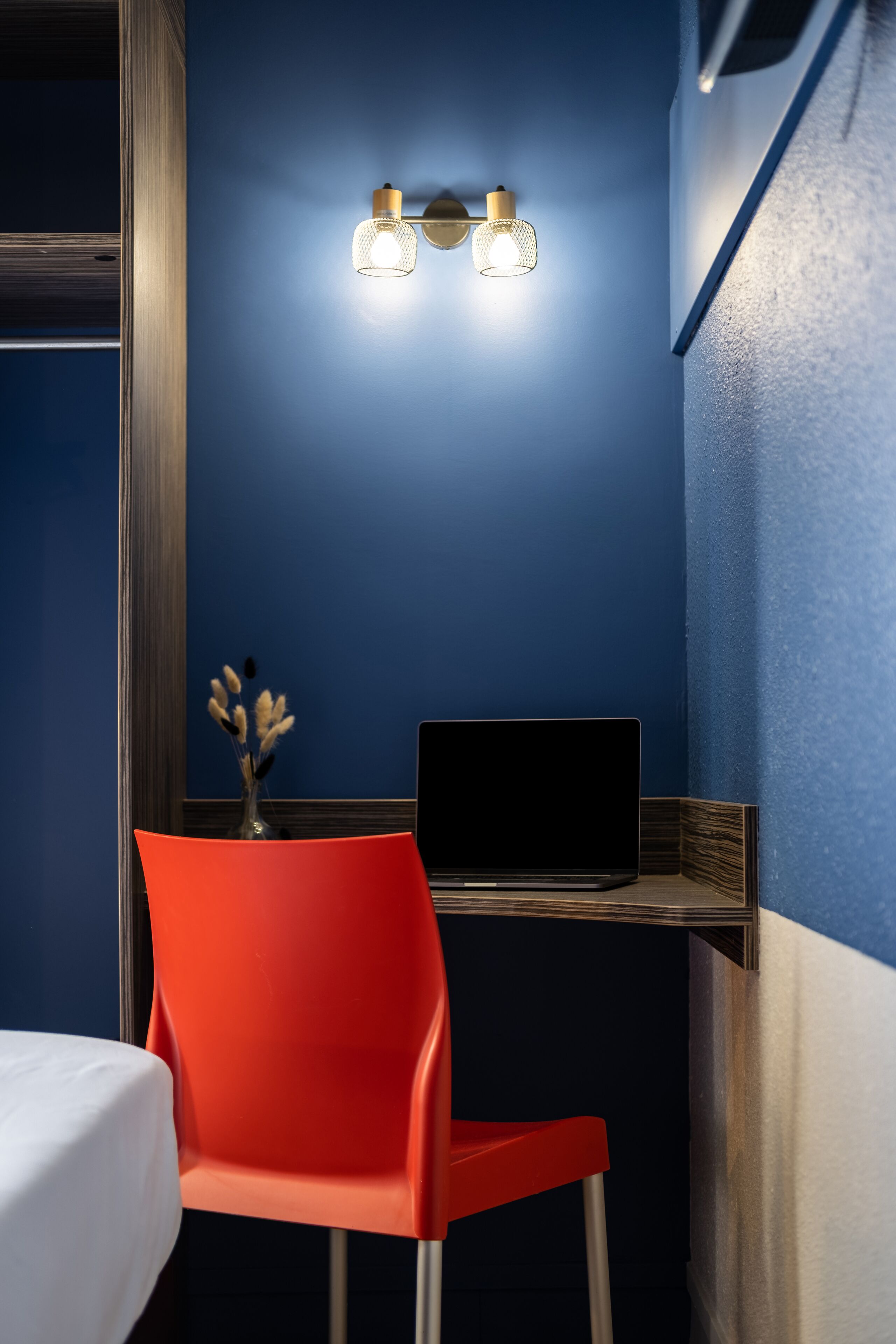 Quarto com parede azul, escrivaninha de madeira, cadeira vermelha, laptop e luminária fixada na parede.