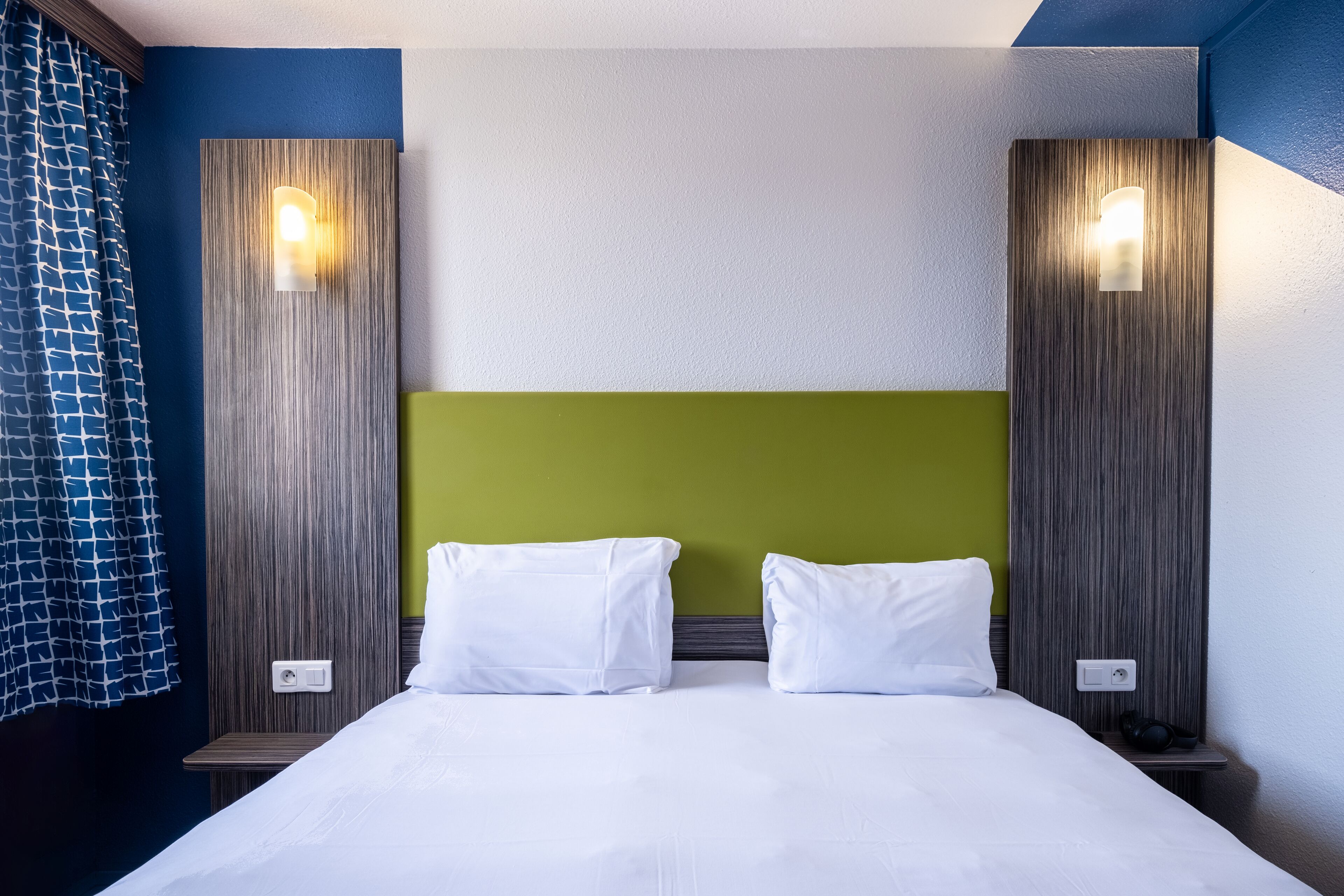 A hotel room with a bed, two bedside tables, a headboard, and wall-mounted lights.