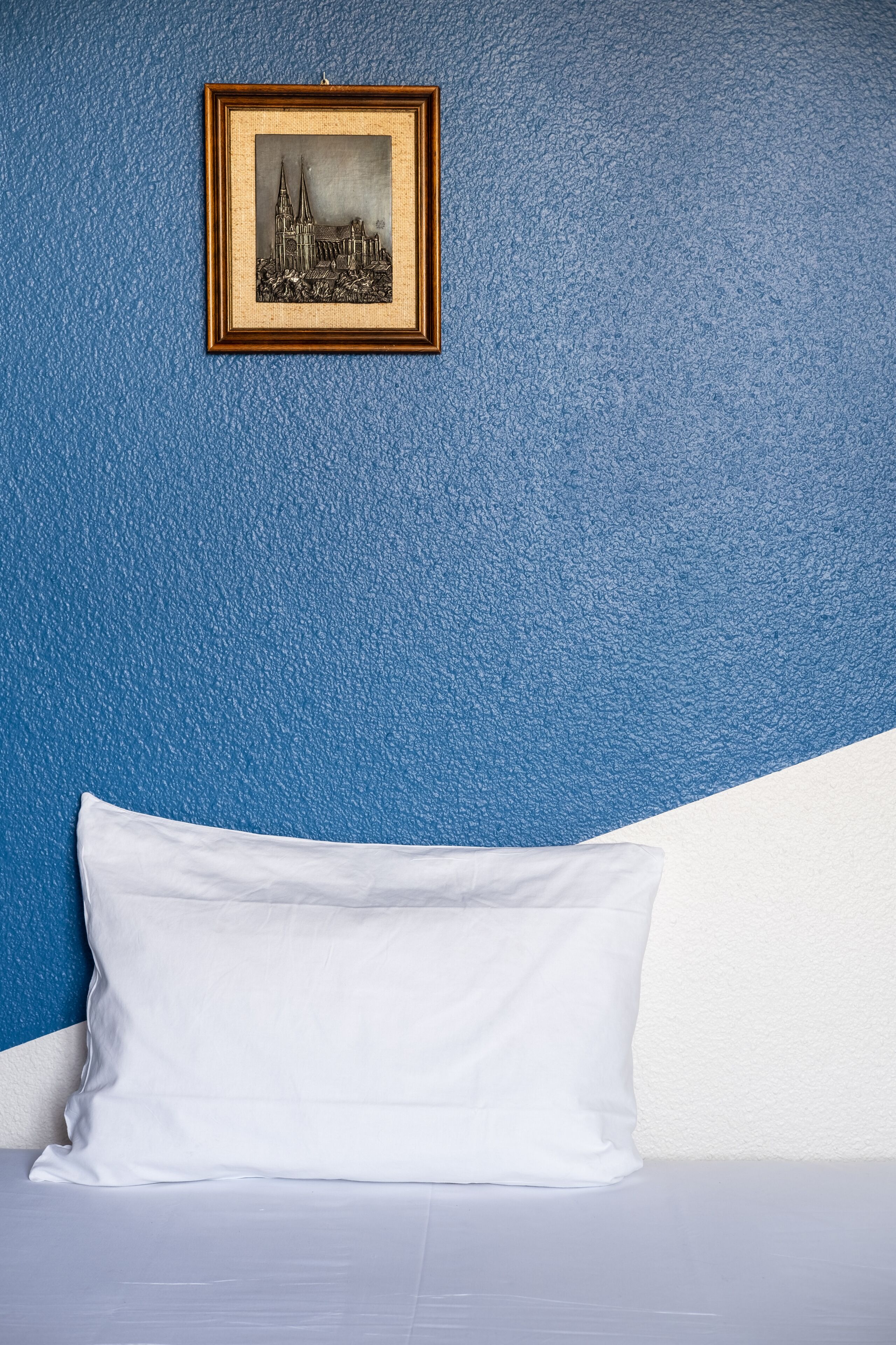 A bed with white bedding against a blue wall, a framed picture of a building above it.