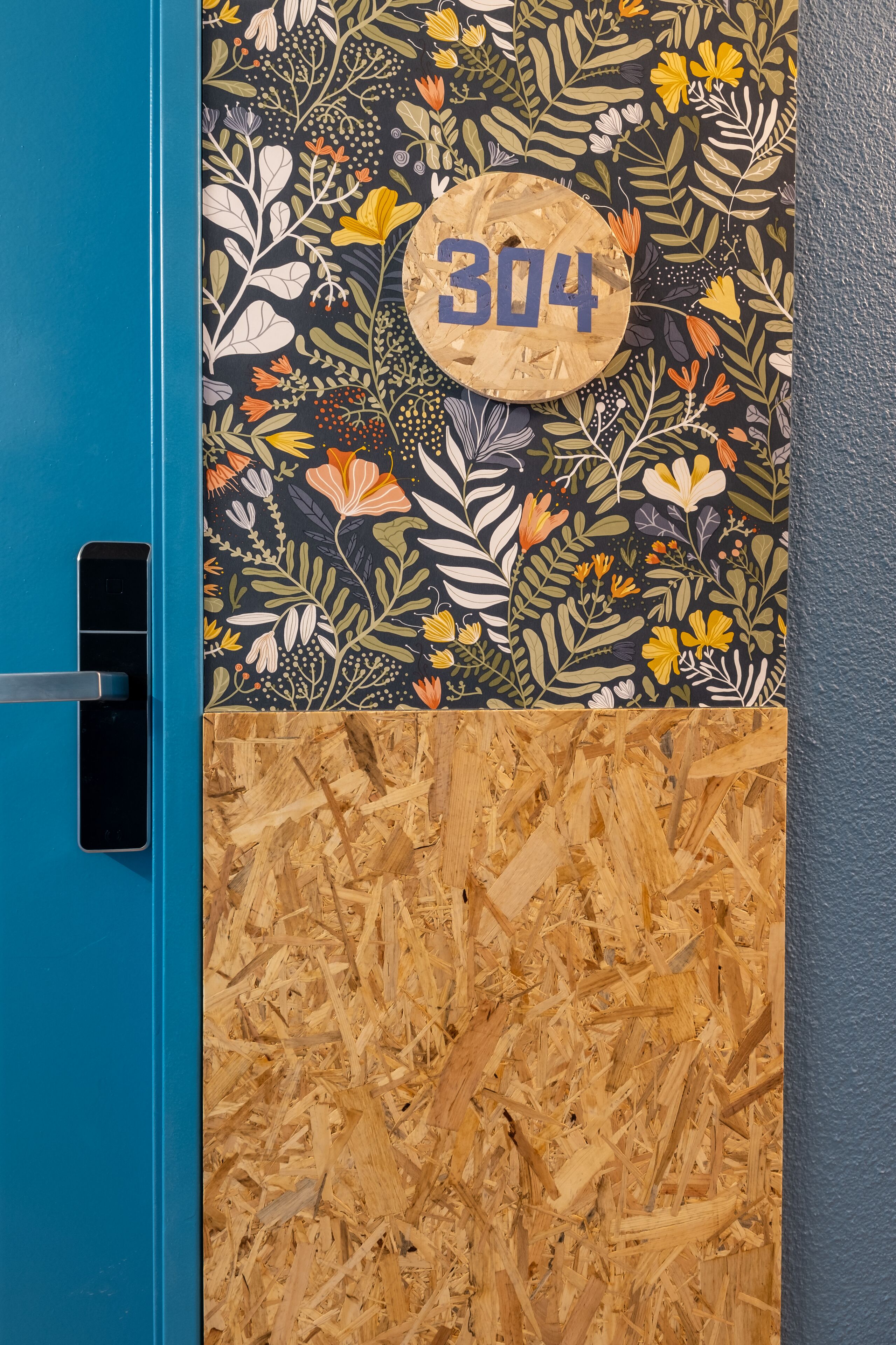 Una puerta azul con cerradura, una puerta de madera con diseño floral y el número 304, y una sección de la puerta cubierta de virutas de madera.
