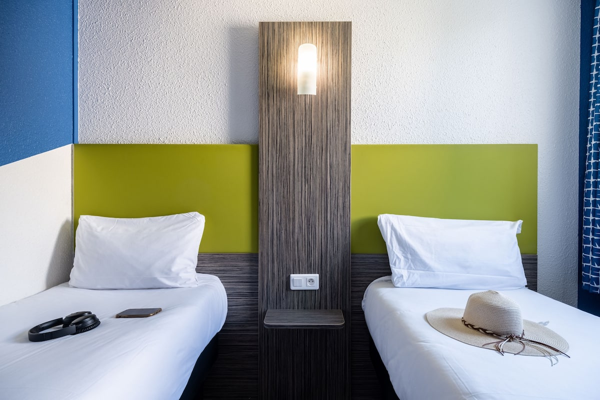 Two single beds with white bedding, a wall-mounted light, and a small shelf.