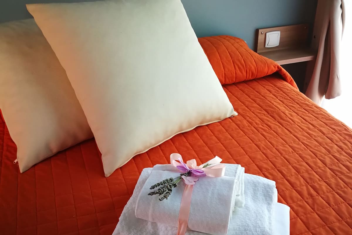 Un lit bien fait, avec une couverture orange, des oreillers blancs et une pile de serviettes pliées, sur laquelle repose une petite branche de lavande.