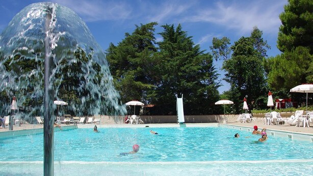 A swimming pool with a water fountain, surrounded by trees and sun umbrellas. - Camping Campo al Fico (Piombino)