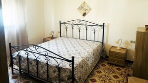 Une chambre à coucher avec un lit en métal, une couvre-lit à motifs, une petite table de chevet avec une lampe, une table de nuit en bois et un tableau accroché au mur.