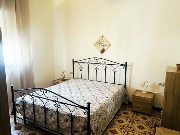 A bedroom with a metal bed frame, patterned bedspread, a small bedside table with a lamp, a wooden nightstand, and a wall-mounted picture.