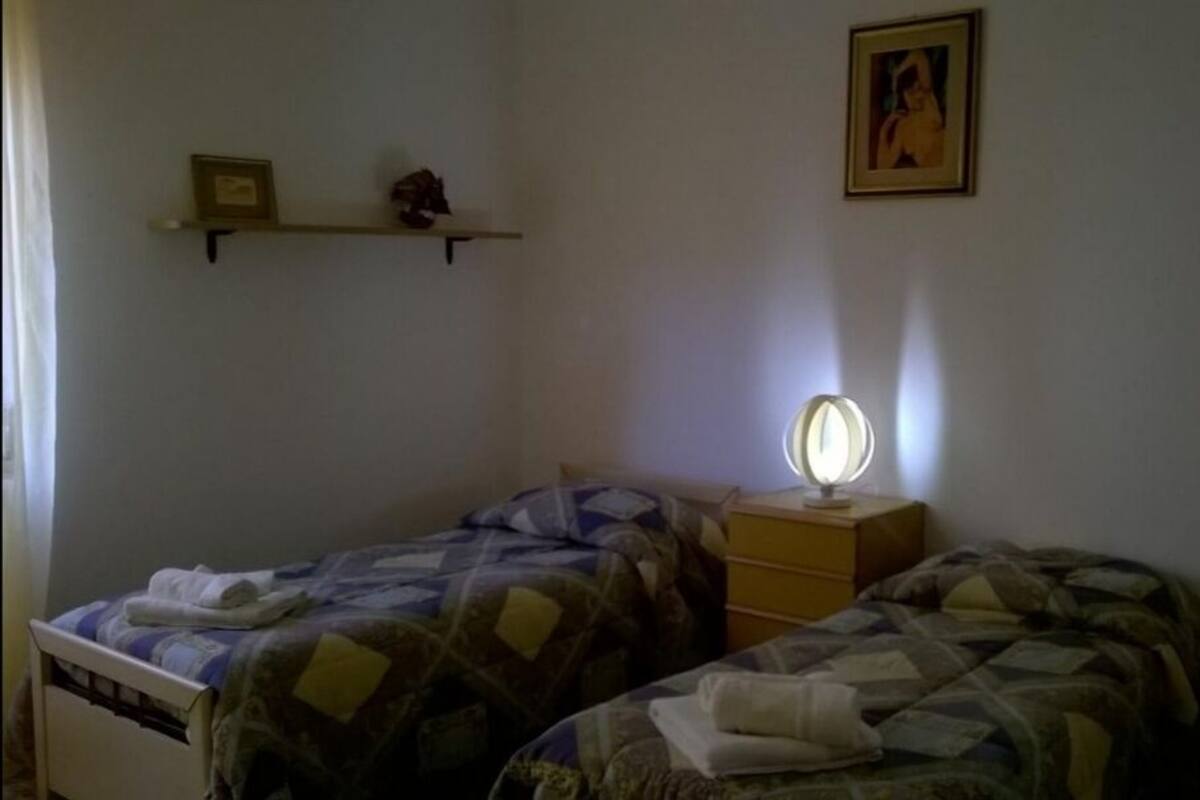 A bedroom with two beds, a nightstand with a lamp, a shelf with a picture, and a wall-mounted picture.