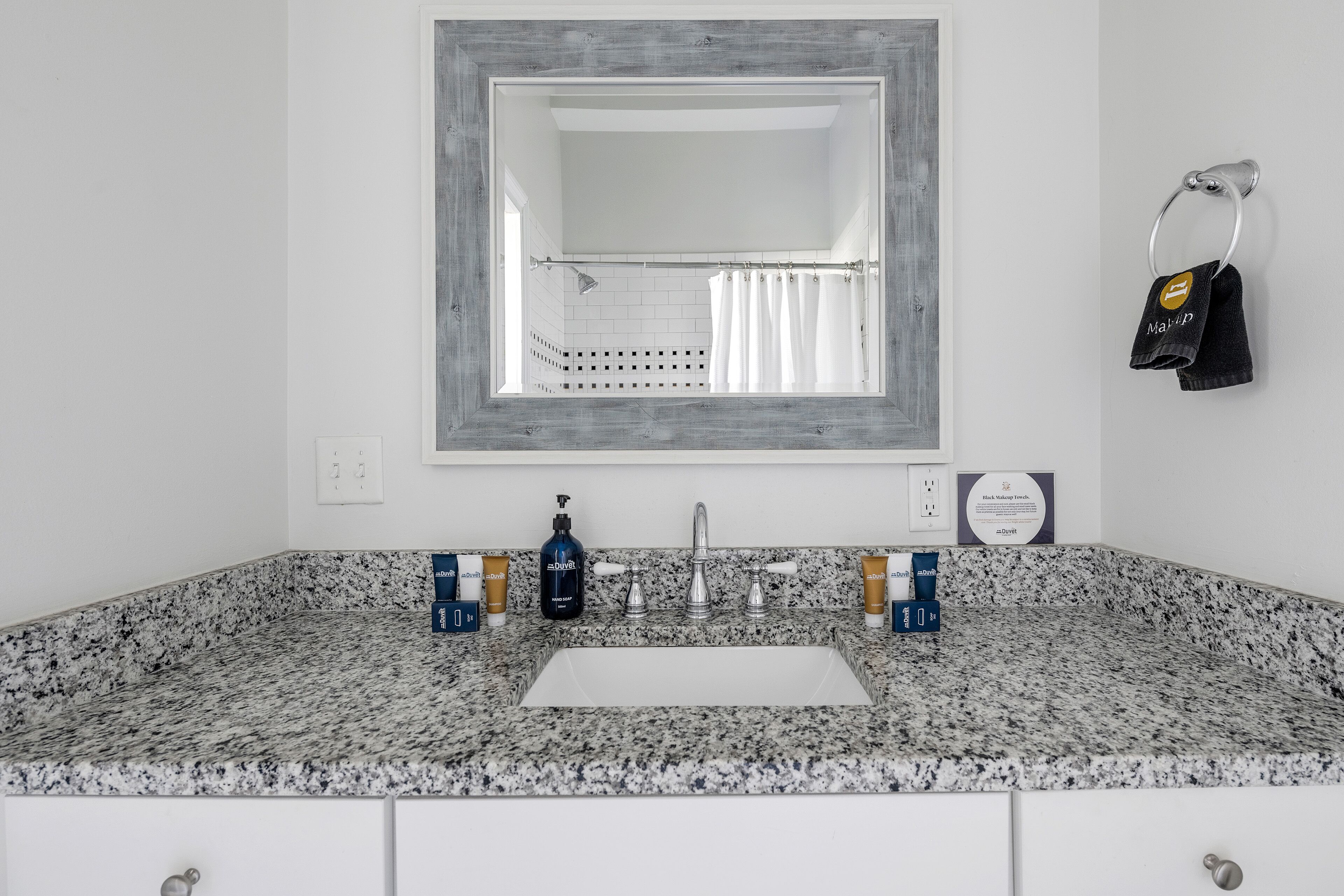 Un baño con encimera de granito, un lavabo blanco y un espejo grande. Hay artículos de aseo en la encimera, y un toallero con una toalla colgada.