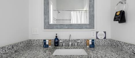Un baño con encimera de granito, un lavabo blanco y un espejo grande. Hay artículos de aseo en la encimera, y un toallero con una toalla colgada.
