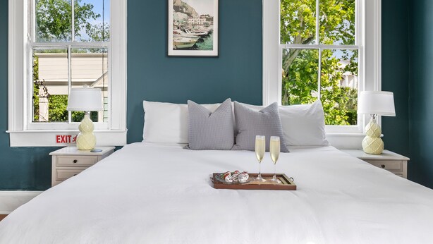 A neatly made bed with a tray of food and two glasses of champagne, a framed picture on the wall, and two windows with a view of greenery and buildings.