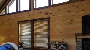 A cozy cabin interior with a wooden ceiling, a fireplace, a black leather sofa, a patterned armchair, and a large window with blinds.