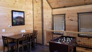 A wooden interior with a foosball table, a dining table with chairs, a painting, and a window with blinds.