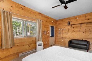 A room with wooden walls, a bed, a ceiling fan, a window with curtains, and a door. - Sunset View | Superior West Twin Lake Home (Lewiston)