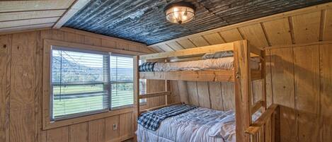 A rustic cabin bedroom with a bunk bed, wooden walls, a large window with a view, and a ceiling light fixture.