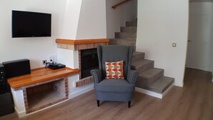 Ein Wohnzimmer mit Kamin, einem grauen Sessel mit gemustertem Kissen, einem Holztisch und einer Treppe im Hintergrund.