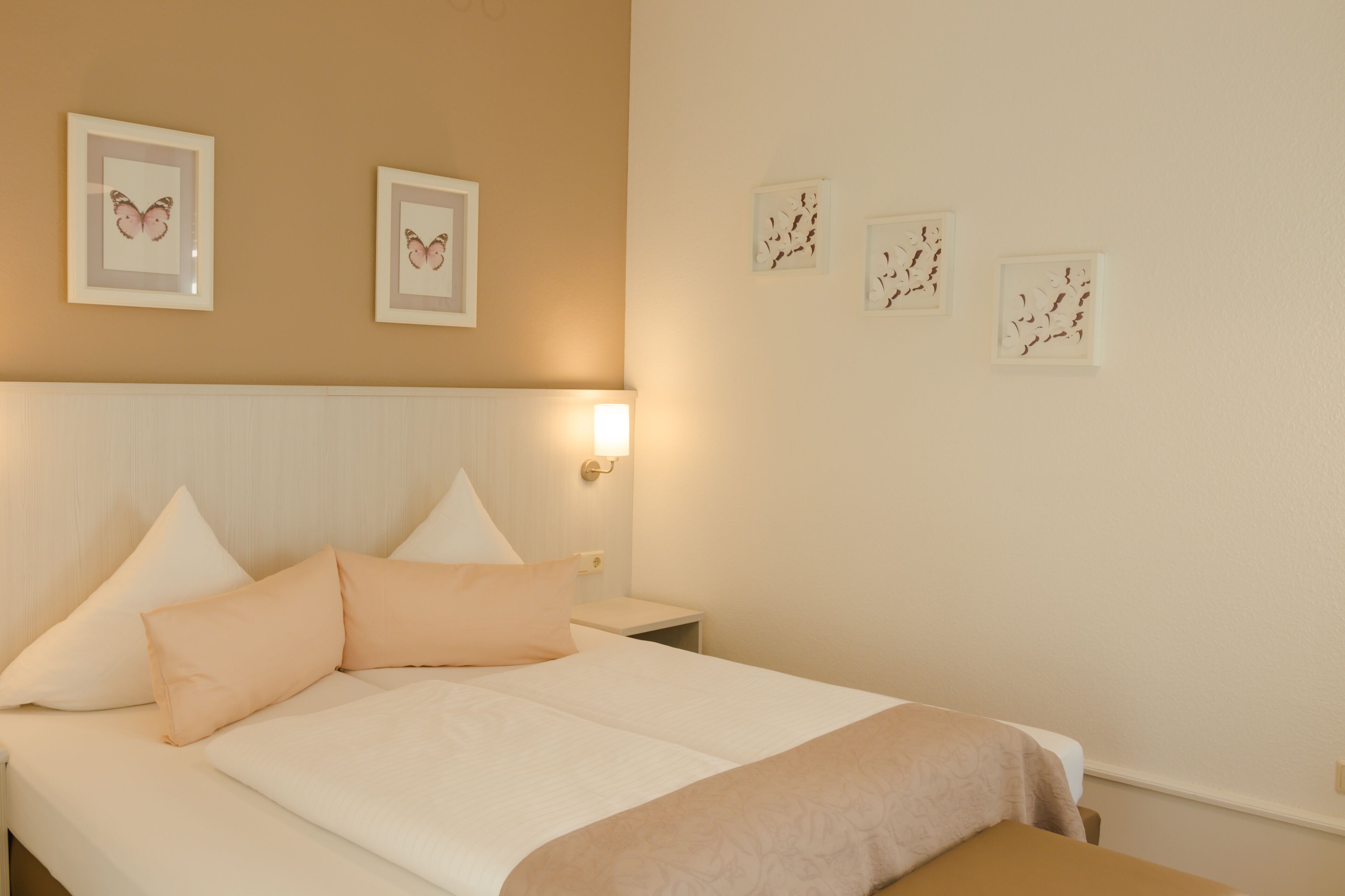 A neatly made bed with a white and peach color scheme, a bedside table, and wall-mounted artwork.