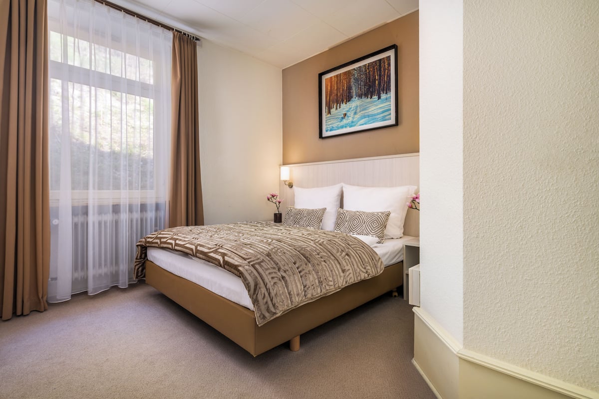 A hotel room with a bed, a painting on the wall, and a window with curtains.