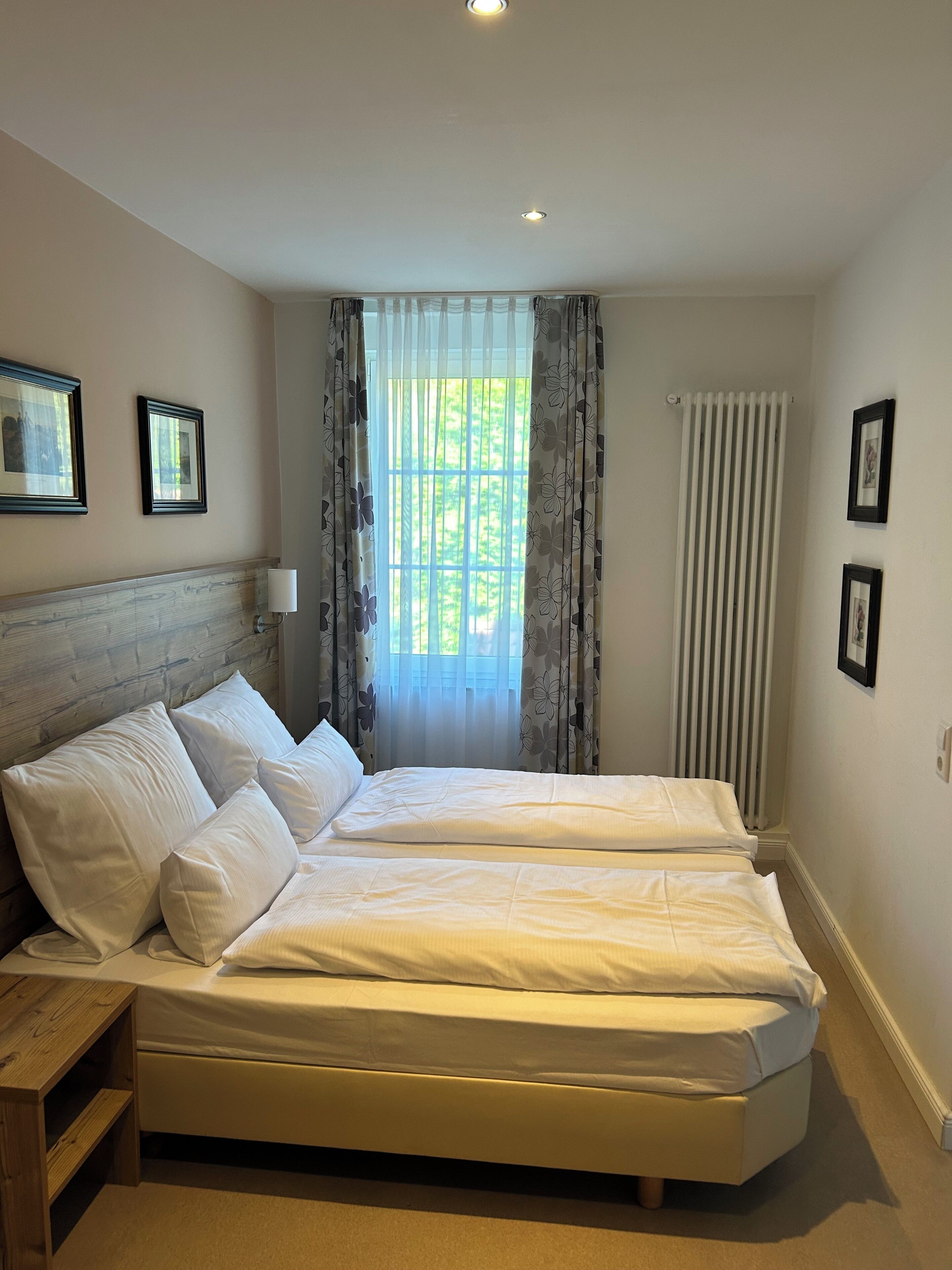 Ein Doppelbett mit weißen Leinentüchern, ein hölzernes Kopfbrett, ein Nachttisch und ein Fenster mit durchsichtigen Vorhängen.