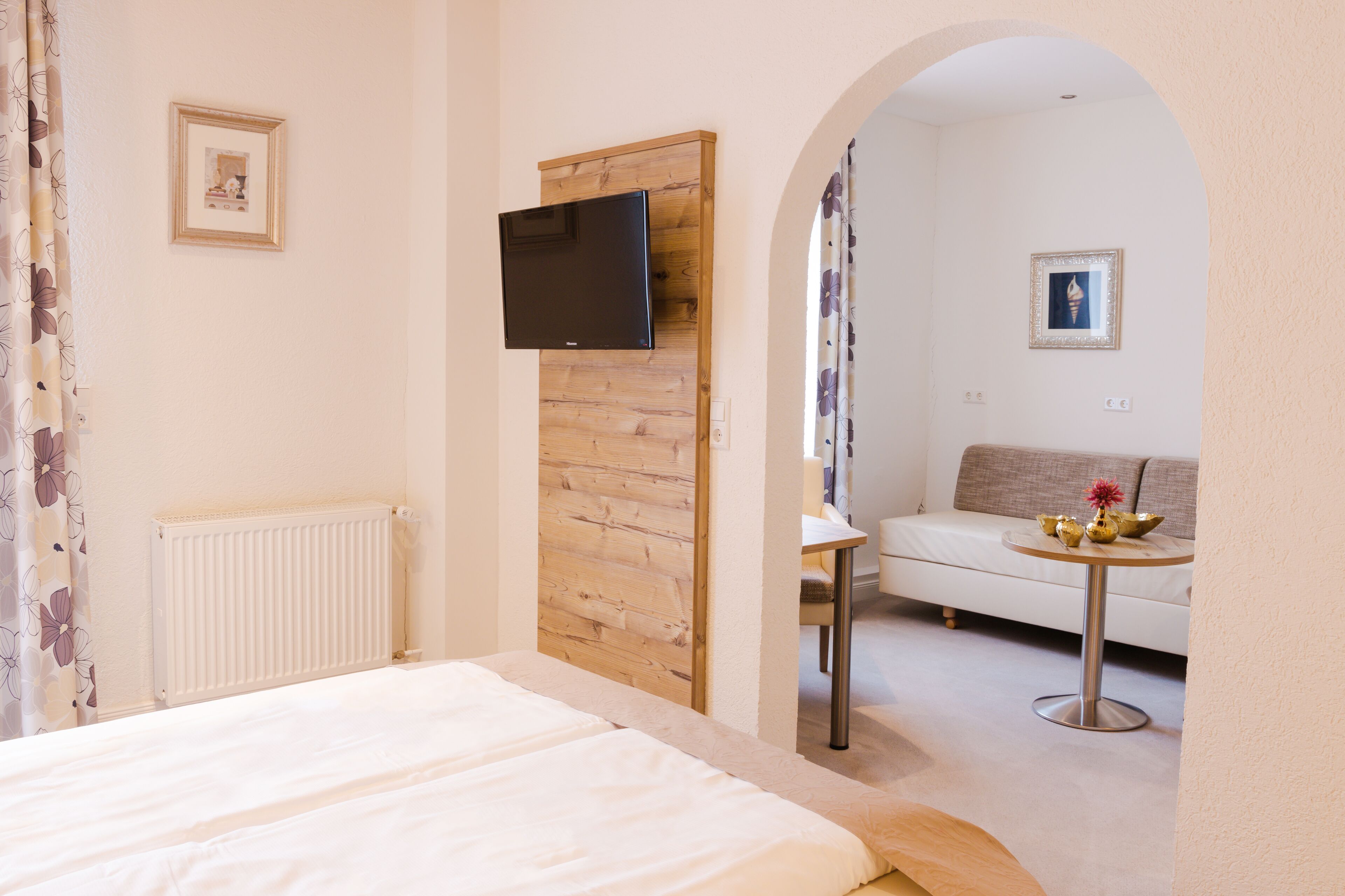 A bedroom with a bed, a TV mounted on a wooden panel, a radiator, and a view of a living area with a sofa and a small table.