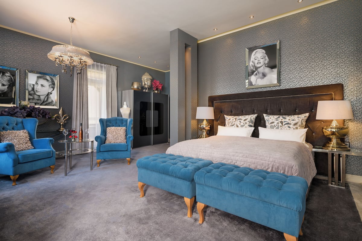 A bedroom with a large bed, two blue armchairs, a chandelier, and a dresser.