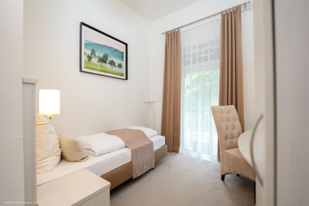 Ein Hotelzimmer mit einem Bett, einem Sessel, einem Nachttisch, einer Lampe und einem Fenster mit Vorhängen.