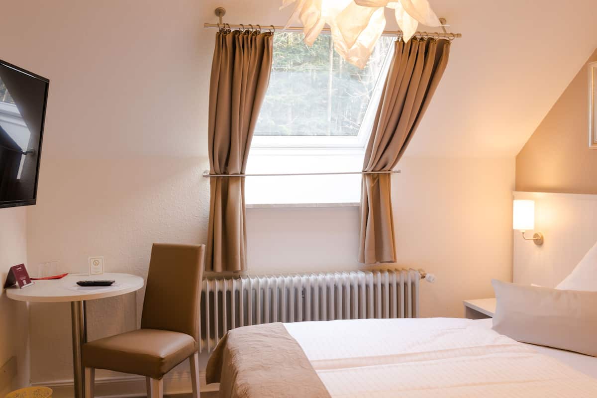 A hotel room with a bed, a chair, a table with a remote control, and a window with curtains.