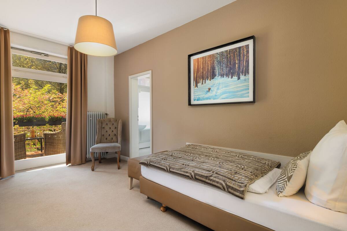 A bedroom with a bed, a chair, a large window, and a framed picture on the wall.