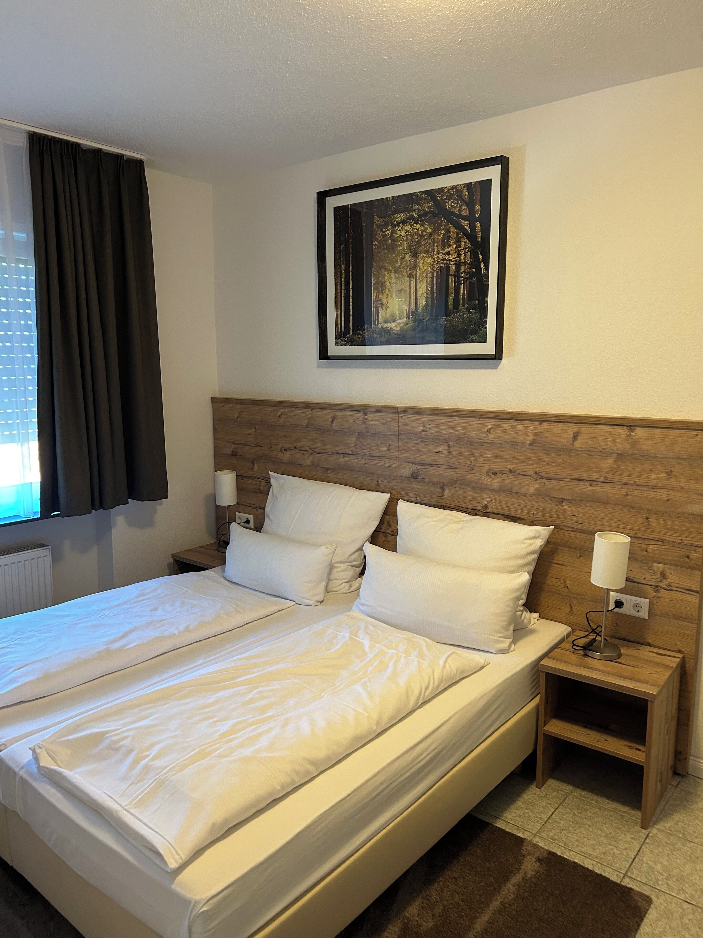 A neatly made bed with white linens, a wooden headboard, a bedside table with a lamp, and a framed picture of a forest on the wall.