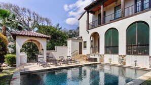 A swimming pool with lounge chairs and a pergola, surrounded by a white building with arched windows and a balcony.